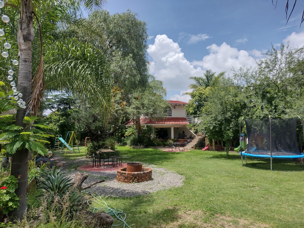 A lush garden is featured, with vibrant greenery surrounding a multi-level building. A fire pit is situated on gravel, while a trampoline and swing set are visible on the lawn. Tall trees provide shade, creating a serene outdoor atmosphere.