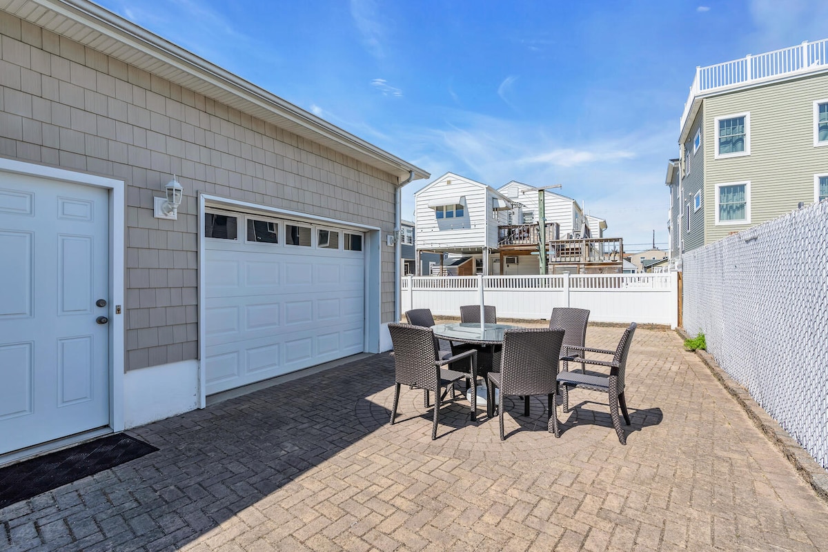 A spacious outdoor patio is displayed, featuring a rectangular table surrounded by four woven chairs. The garage door is positioned to the left, and a white fence adds privacy to the area. Nearby homes are visible in the background, enhancing the neighborhood feel.