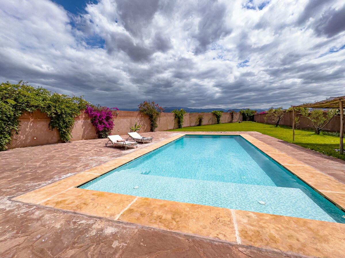 The outdoor pool is surrounded by lush greenery and vibrant flowering plants. Two lounge chairs are positioned next to the pool, offering a space for relaxation. A clear sky with scattered clouds creates a serene atmosphere over the tranquil waters.