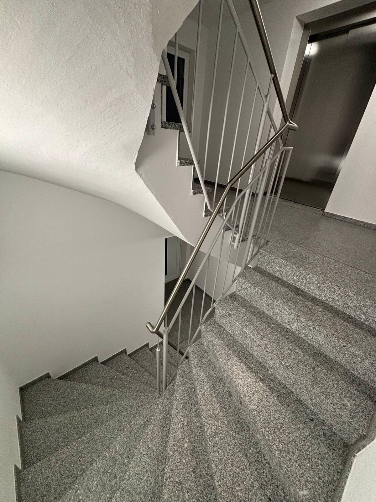 A spiral staircase is depicted, featuring grey stone steps and metallic railing. Natural light is visible in the upper area, highlighting the clean lines and neutral tones of the surrounding walls. An elevator door is positioned on the right side of the image.