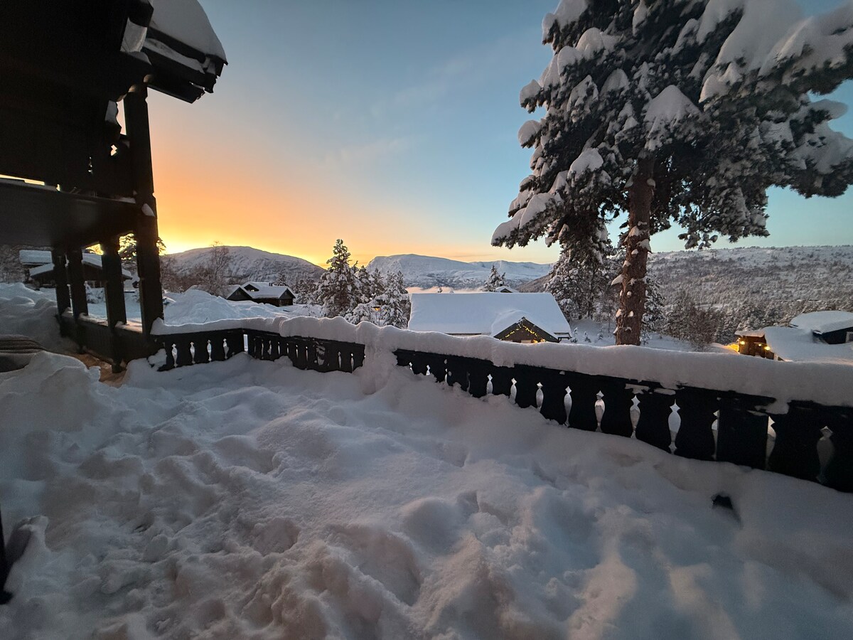 A winter scene captures a snowy terrace, framed by wooden railing, as the golden hues of sunset illuminate the distant mountains. Fresh snow blankets the ground, creating a serene atmosphere. The landscape reveals an inviting view of neighboring cabins amidst the tranquil snow-covered surroundings.