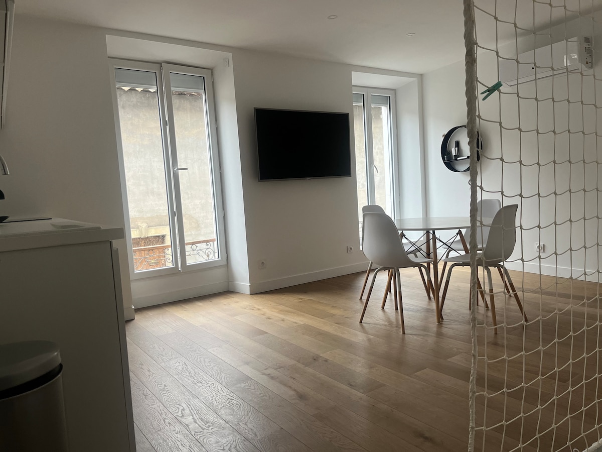 The image showcases a bright and open living area, featuring two large windows allowing natural light. A modern dining table with white chairs is centered in the room, while a flat-screen television is mounted on the wall opposite. The wooden flooring adds warmth to the space.