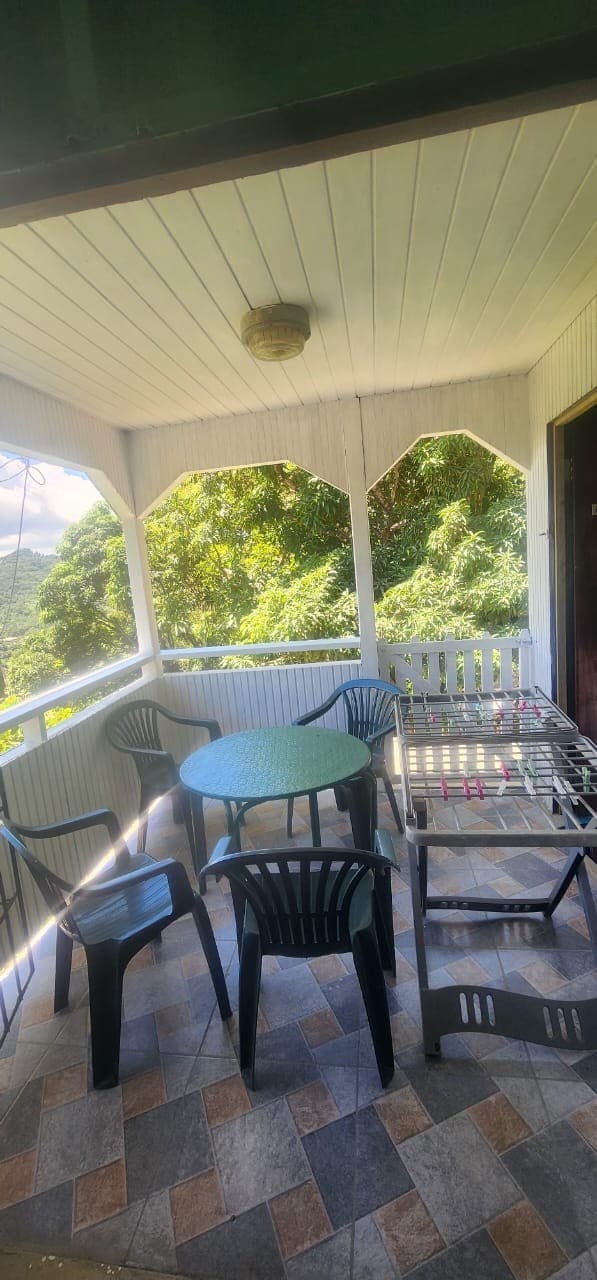 A covered outdoor seating area is shown, featuring a round table surrounded by four black chairs. A drying rack is positioned nearby, with tiled flooring that complements the natural scenery visible through the open space. Lush greenery is present in the background.