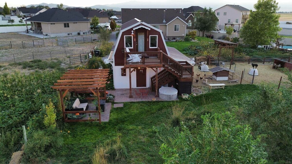 An aerial view of a rustic barn with a deck and patio area, surrounded by green grass and garden patches. The barn features a sloped roof and wooden accents, while outdoor seating areas are visible alongside the garden. Neighbors' homes are seen in the background.