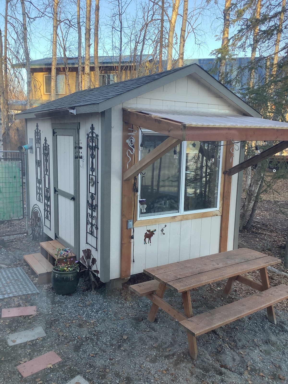 A small, charming structure is depicted with a wooden porch and large window featuring decorative accents. A picnic table is positioned nearby, surrounded by gravel and a few plants. The rustic design is complemented by the natural setting of trees in the background.