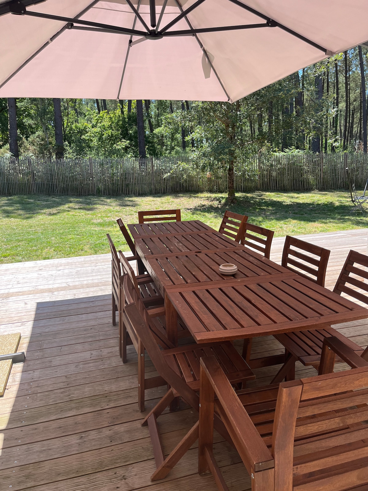 An outdoor dining area is featured, showcasing a large wooden table surrounded by matching chairs. A light-colored umbrella provides shade over the table, while a grassy area and a natural wooden fence are visible in the background, enhancing the outdoor setting.