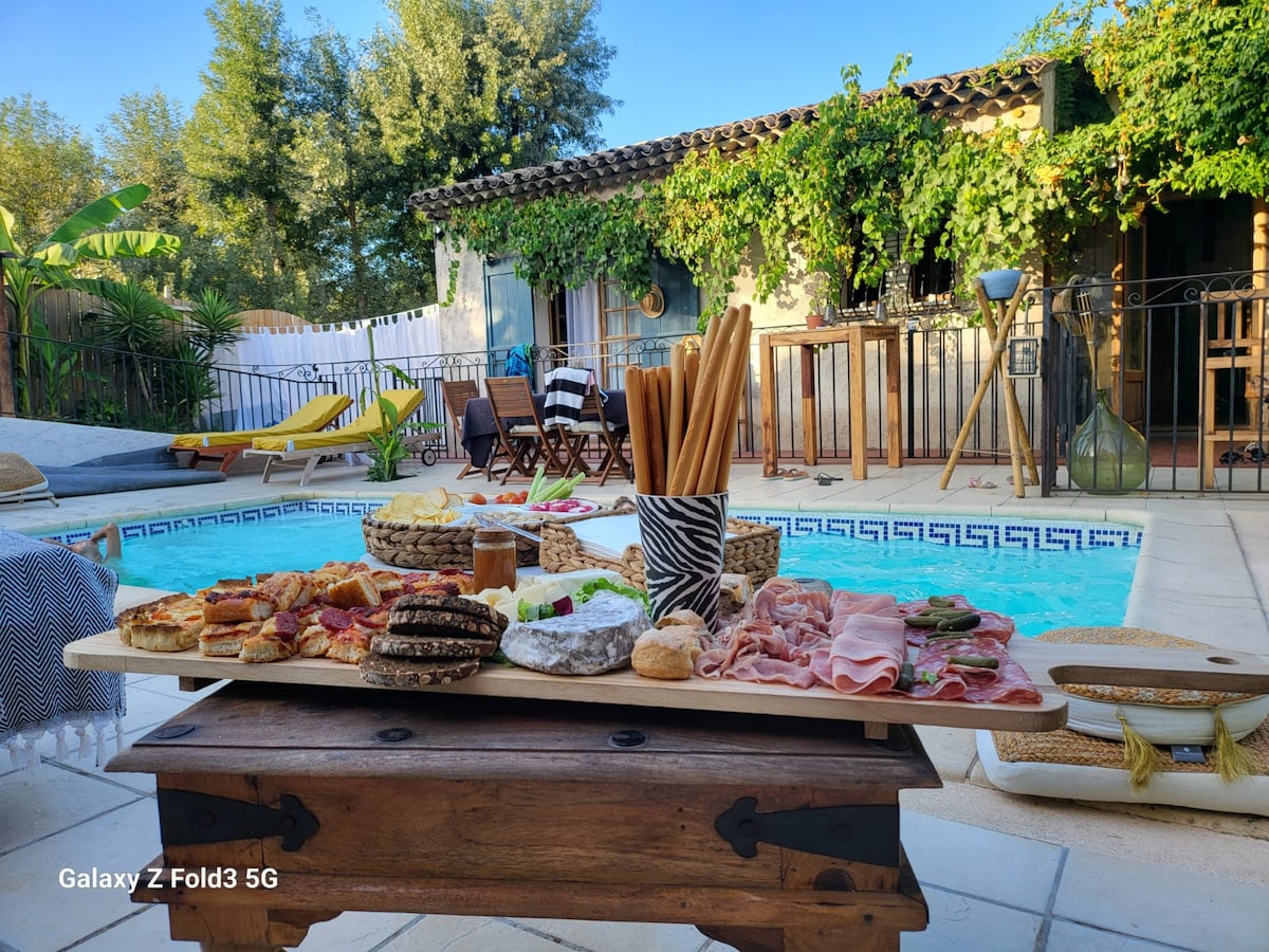 A beautifully arranged outdoor dining area is presented by a sparkling pool. Various platters feature an assortment of savory snacks and breads on a wooden table. Sun loungers and chairs surround the pool, with lush greenery enhancing the tranquil atmosphere.
