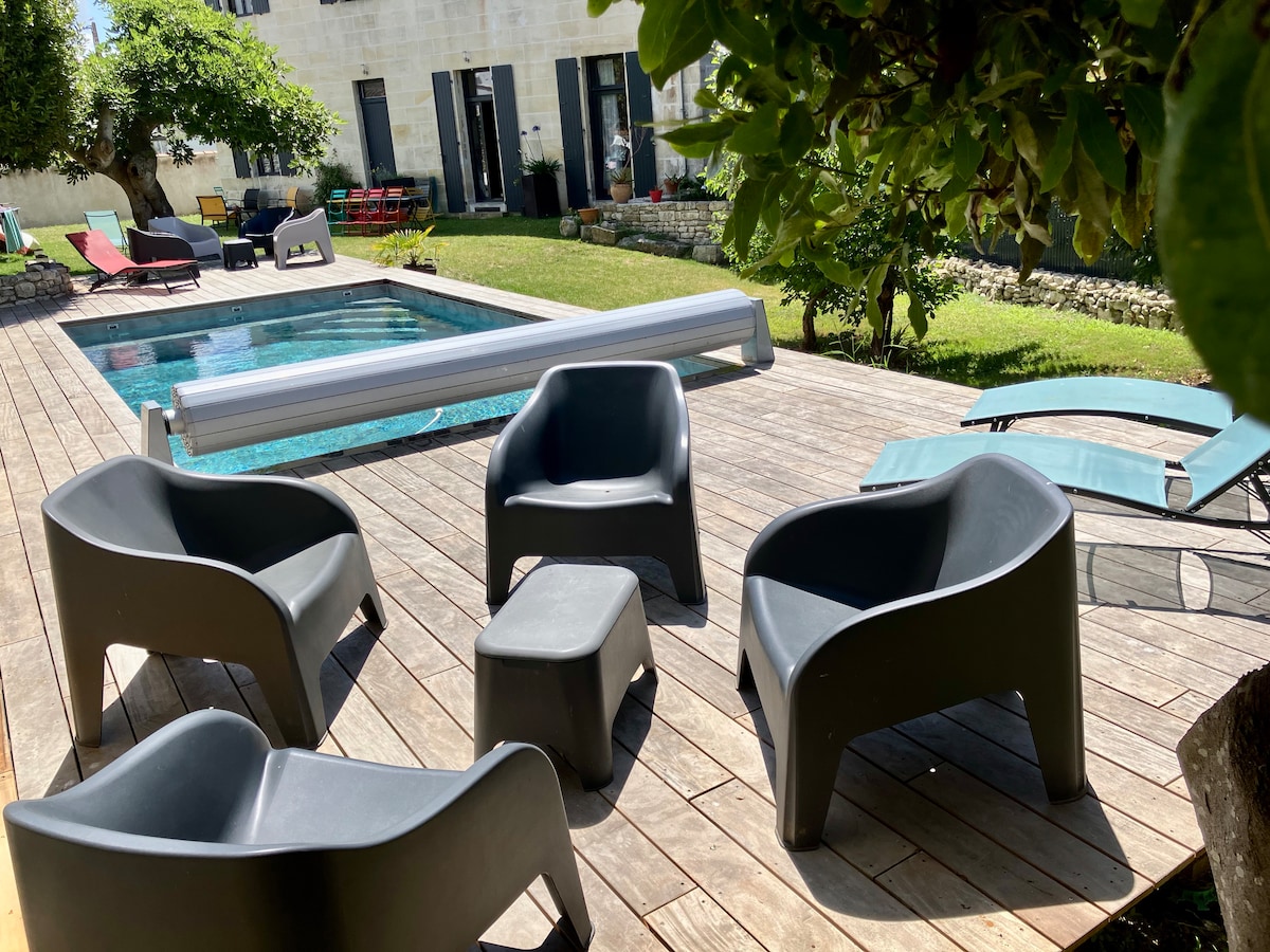 A wooden deck area features a heated swimming pool surrounded by modern, dark grey lounge furniture. Four ergonomic chairs and a small side table are arranged in a social setting, while sun loungers are positioned nearby. Lush greenery provides a natural backdrop.