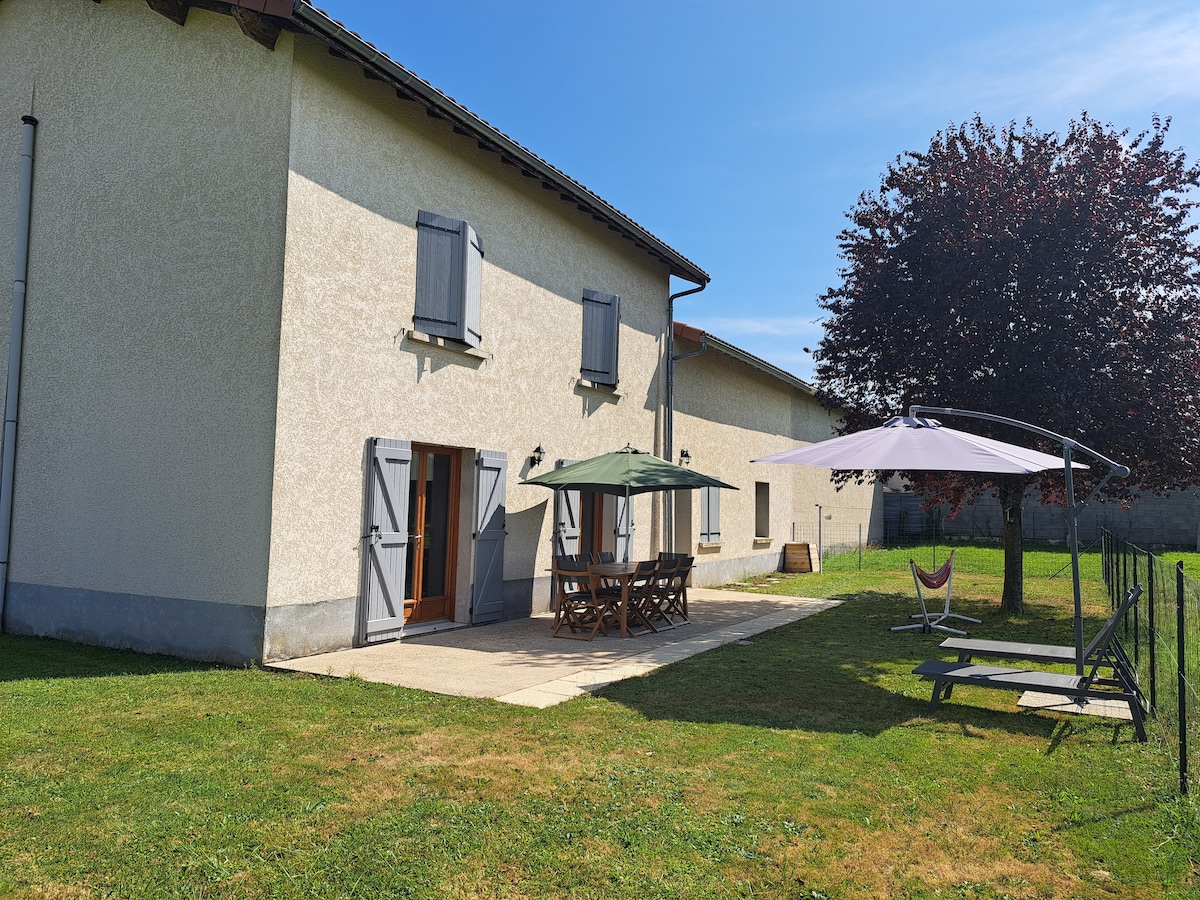 The exterior of the house is visible, showcasing a spacious patio with a large wooden dining table and umbrellas for shade. A tree provides additional greenery, while a hammock and seating area enhance the outdoor space. The sky is clear and blue, creating a bright, inviting atmosphere.
