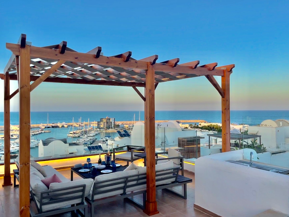 A rooftop terrace features a wooden pergola, providing shade over comfortable seating. The area offers panoramic views of the sea and harbor. A table is set for refreshments, enhancing the inviting outdoor space.