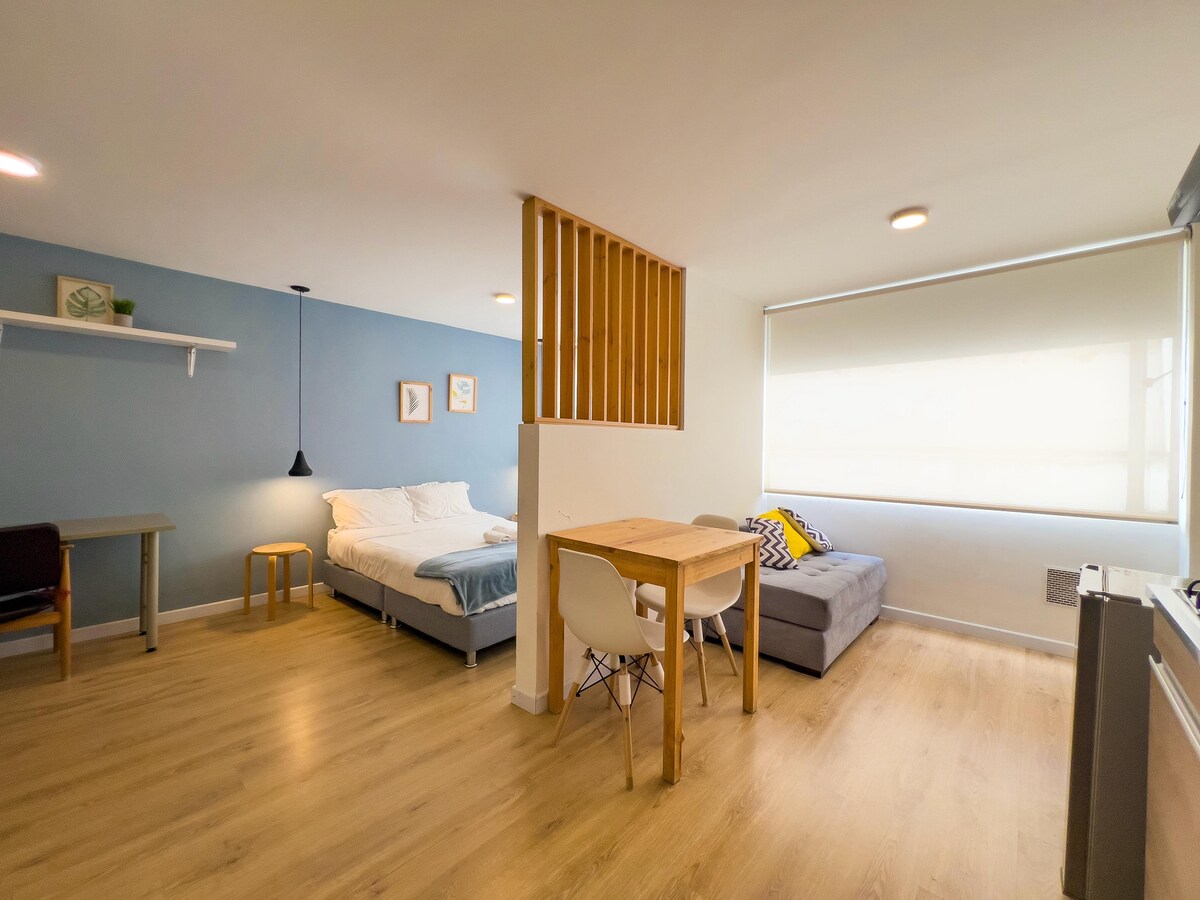 An open and functional living space features a spacious bed and a comfortable gray sofa. A small dining table with chairs is positioned near a window that allows natural light. A softly separated sleeping area enhances the overall layout, with minimalist decor present throughout.