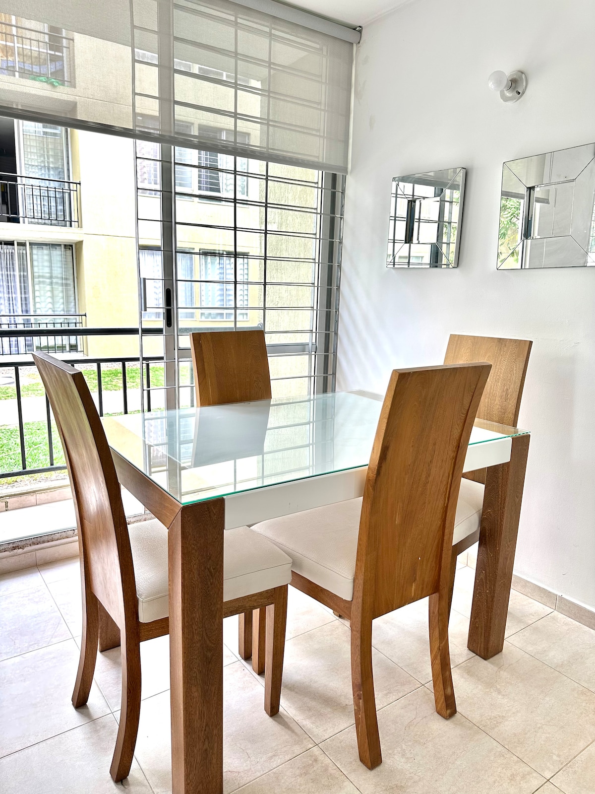A dining area features a glass-top table surrounded by four wooden chairs. Natural light flows through the large window with sliding panels, illuminating the space and highlighting the adjacent greenery visible outside.