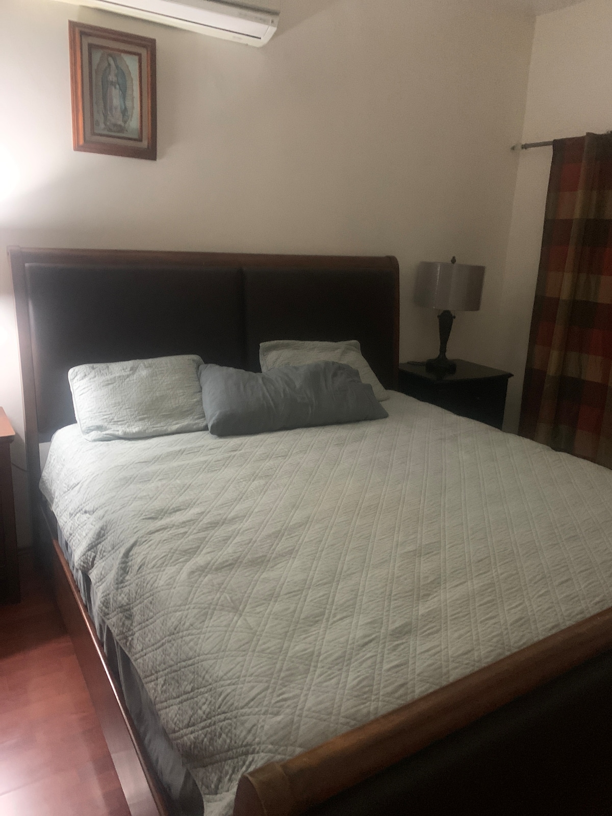 A spacious bed with a quilted coverlet is positioned against a wall, complemented by decorative pillows. A lamp on a side table provides soft lighting, while a patterned curtain adds a touch of color. A framed artwork is displayed above the bed.