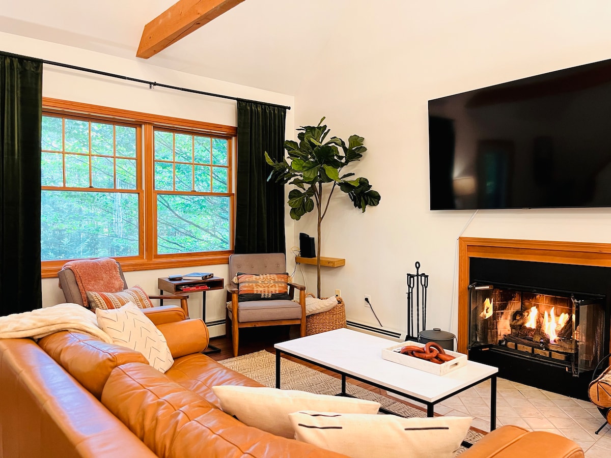 A cozy living area features a large leather sectional sofa facing a modern fireplace. A flat-screen television is mounted on the wall above a wooden mantel. Natural light enters through a pair of large windows, complemented by dark curtains and a decorative indoor plant.