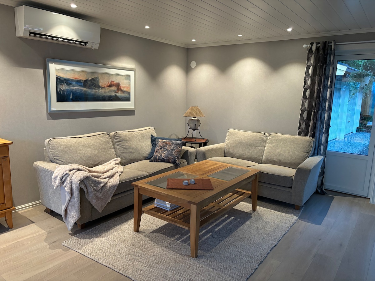 A well-lit living area features a cozy grey sofa set with plush cushions and a decorative throw. A wooden coffee table sits at the center, complemented by a small lamp. Light-colored walls and a soft rug enhance the room's inviting feel.
