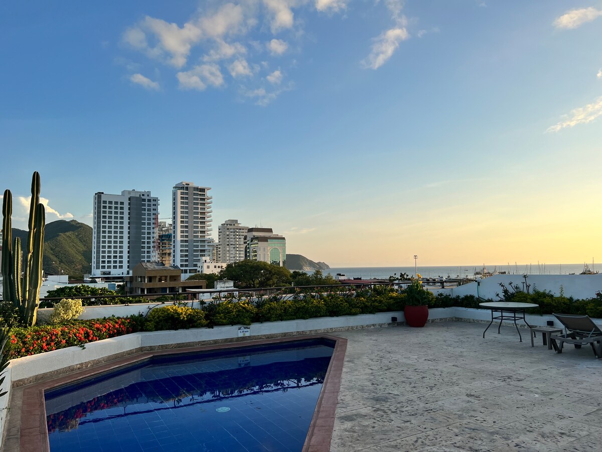 A serene rooftop space features a tiled pool surrounded by landscaped areas with flowering plants. The view includes a skyline of modern buildings, hills in the background, and a glimpse of the ocean under a clear sky.