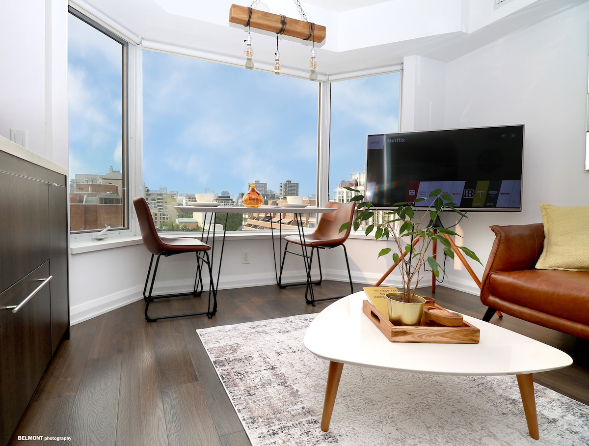 A bright living space is highlighted by large windows offering an expansive view of the city. A modern dining table is paired with stylish chairs, while a cozy seating area features a sleek sofa. A flat-screen TV is mounted on the wall, and a decorative plant adds a touch of nature.