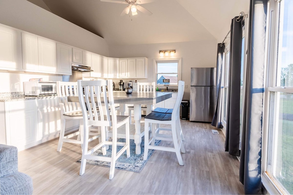 A well-lit kitchen showcases modern white cabinetry and stainless appliances. A spacious island with high chairs invites gatherings. Natural light streams through large windows, revealing views of the outdoors. Neutral-toned flooring and walls contribute to a clean, open atmosphere.