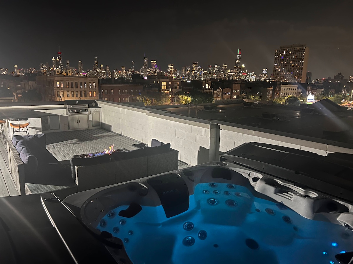 A rooftop space features a hot tub illuminated in blue, accompanied by a seating area with a sofa. A city skyline stretches in the background, showcasing a vibrant urban landscape against the night sky.