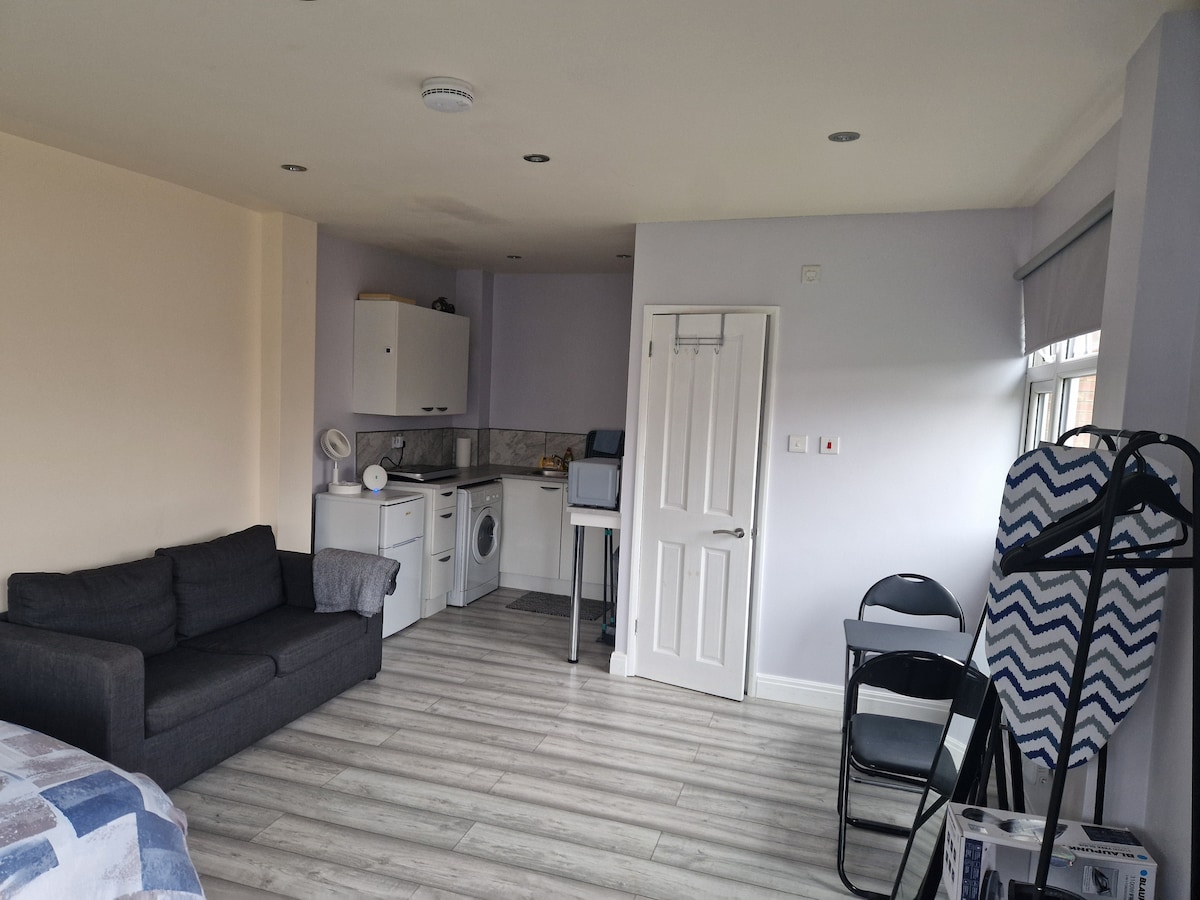 An open-plan living area features a comfortable sofa, a kitchenette equipped with essential appliances, and a dining space. Light-colored walls and flooring enhance the spacious feel. A wardrobe and two folding chairs are also present, providing practical amenities for guests.