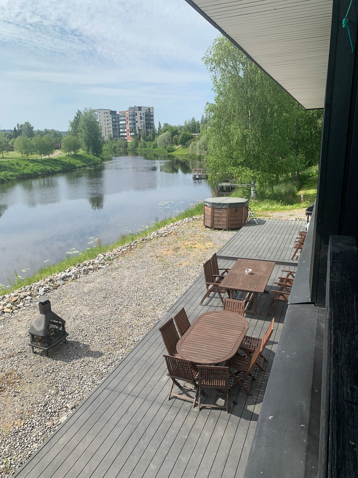 A spacious wooden deck extends along the riverbank, furnished with multiple tables and chairs. A small sauna structure is visible nearby, while lush greenery surrounds the area. The calm water reflects the clear sky, providing a serene outdoor space for gatherings.