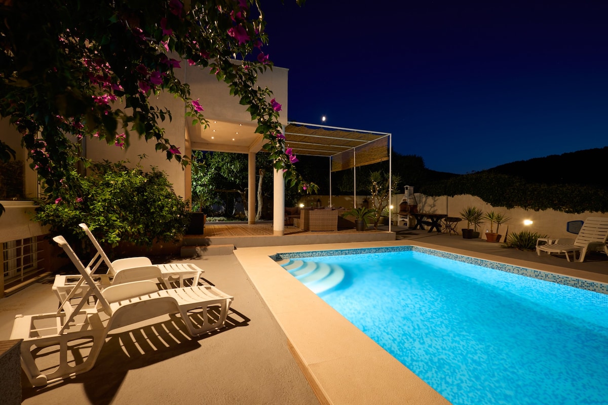 A well-lit swimming pool glimmers under a night sky, surrounded by lush greenery. Loungers are positioned nearby, offering space for relaxation. The outdoor seating area features a protective canopy, creating a cozy environment for gatherings.