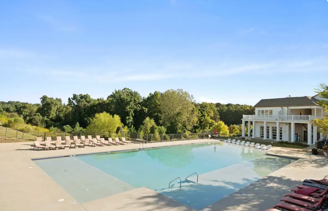 A large outdoor pool is surrounded by lounge chairs and offers unobstructed views of lush greenery. The pool features stairs for easy entry and a spacious deck area. A two-story building is visible in the background, contributing to the inviting atmosphere.