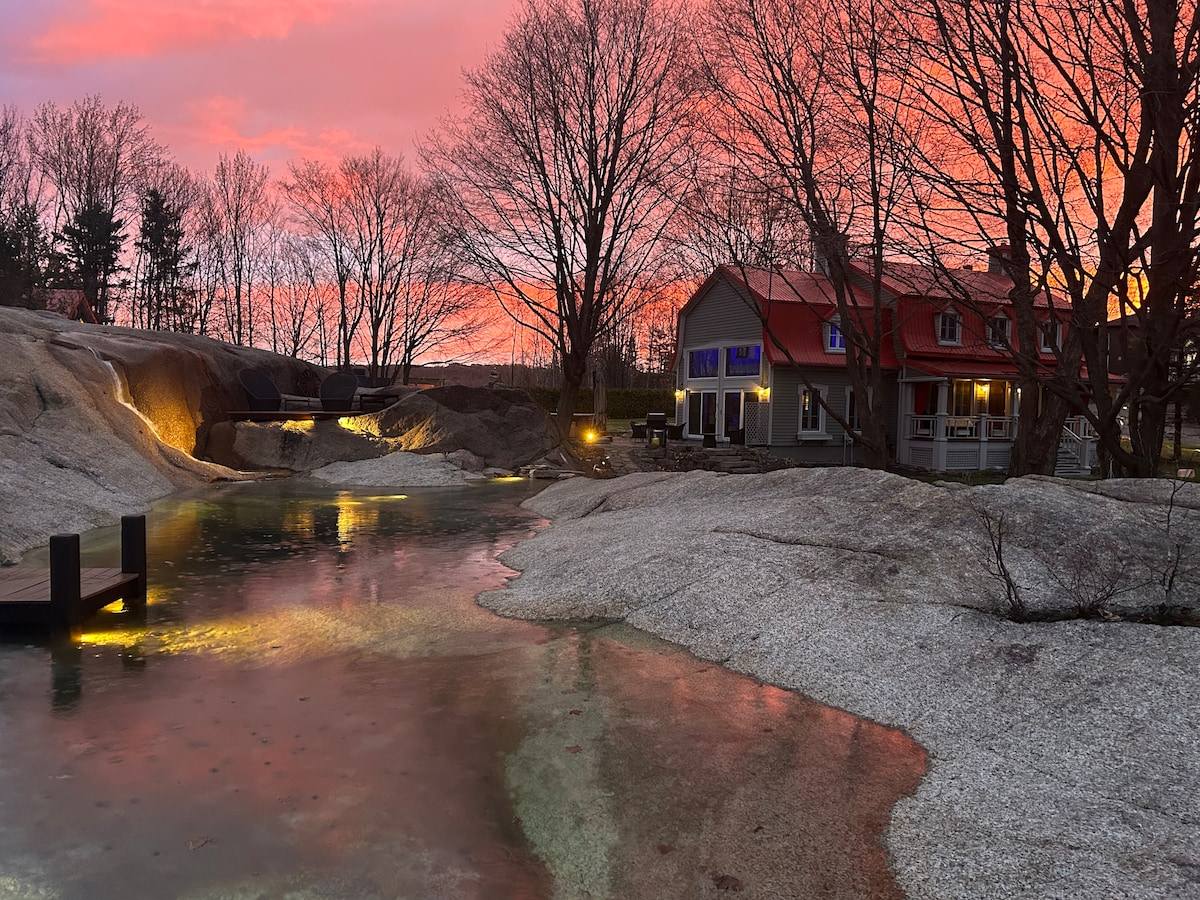 A natural estate is set against a vibrant sunset, with soft pink and orange hues reflecting off a gracefully shaped swimming pool designed in granite. Surrounding trees are silhouetted, and a charming house with a red roof is positioned nearby, enhancing the serene landscape.