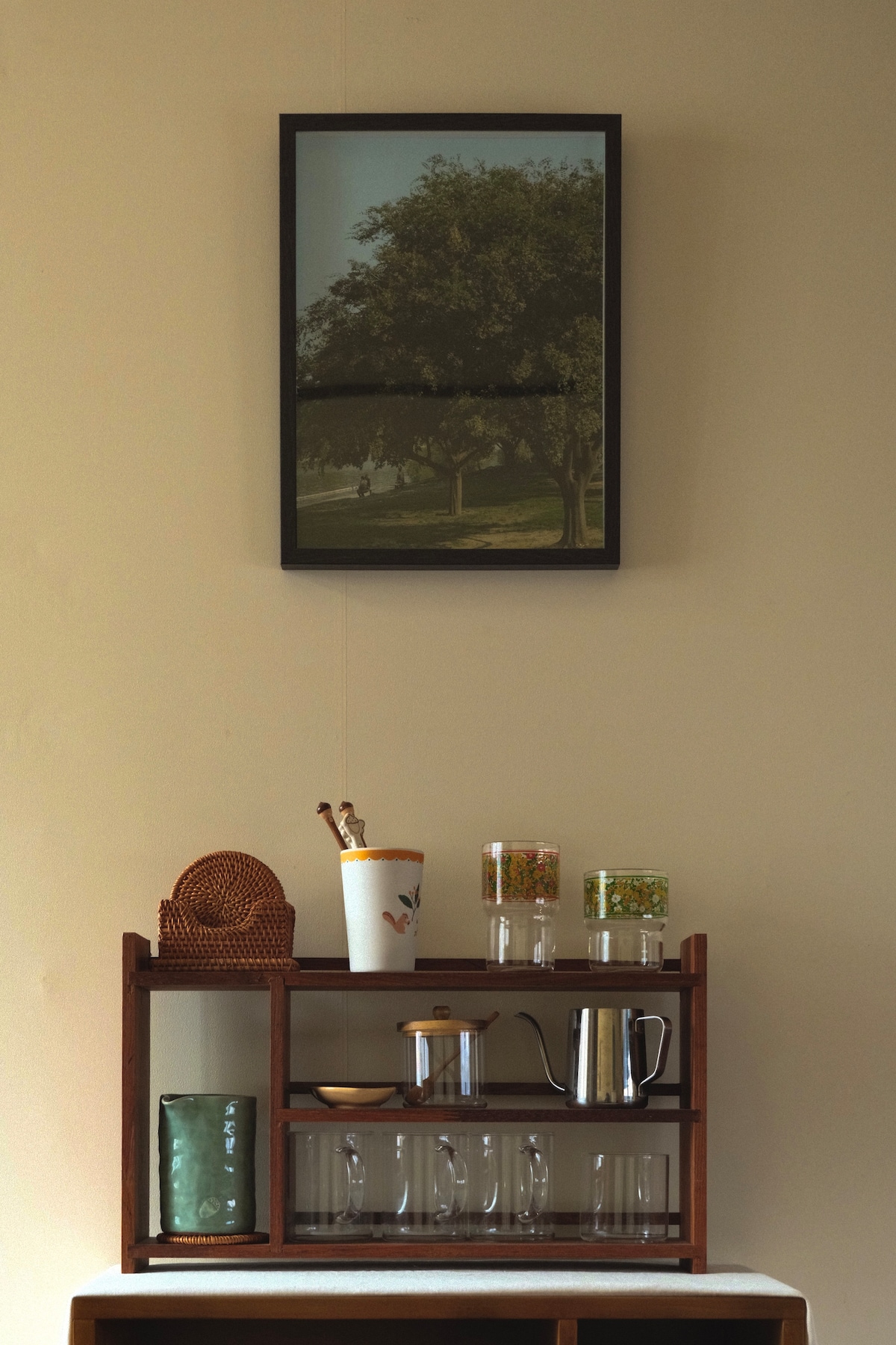 A wooden shelf holds glass cups, a small kettle, and jars filled with various items. Above, a framed artwork depicting a tree and landscape is mounted on a neutral wall, complementing the serene space.