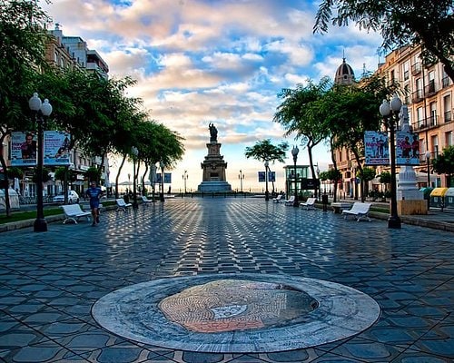 A wide plaza is depicted with a central monument surrounded by pedestrian pathways. Lush trees line the sides, offering shade. Several benches are positioned for seating, while banners and street signs provide information. The cloudy sky creates a soft light over the area.