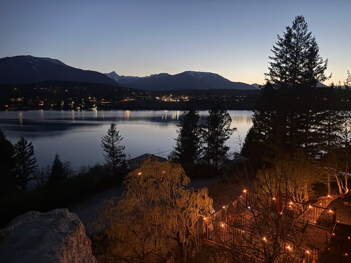 A serene view of Windermere Lake is captured at twilight, reflecting the soft glow of city lights on the water's surface. Majestic mountains are seen in the background, framed by trees. Warm lights illuminate the surrounding area, creating a calm ambiance.