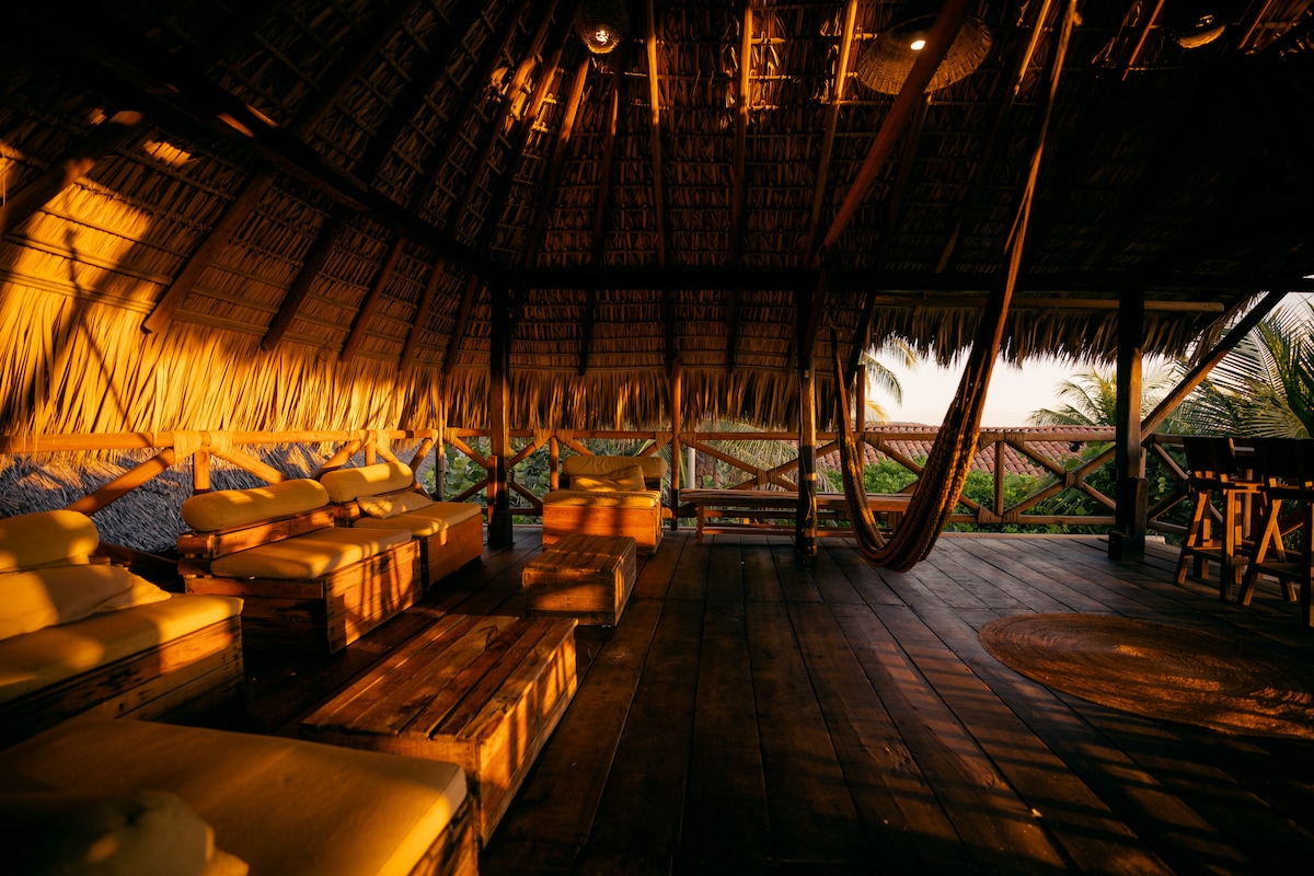 A spacious common area is highlighted by a thatched roof, with natural light pouring in during sunset. Comfortable seating arranged on wooden platforms is seen, alongside hanging hammocks that create a relaxed atmosphere. A round rug complements the wooden flooring, enhancing the inviting environment.