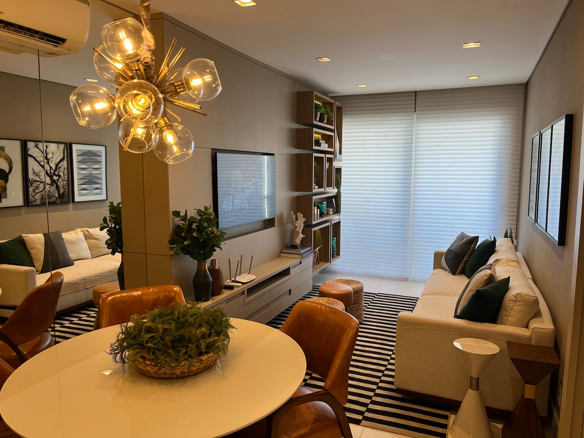 A modern living area is presented, featuring a light-colored sofa alongside a wooden shelving unit. A round dining table with stylish chairs is positioned in the foreground. Natural light enters through large windows with sheer blinds, creating a warm atmosphere.