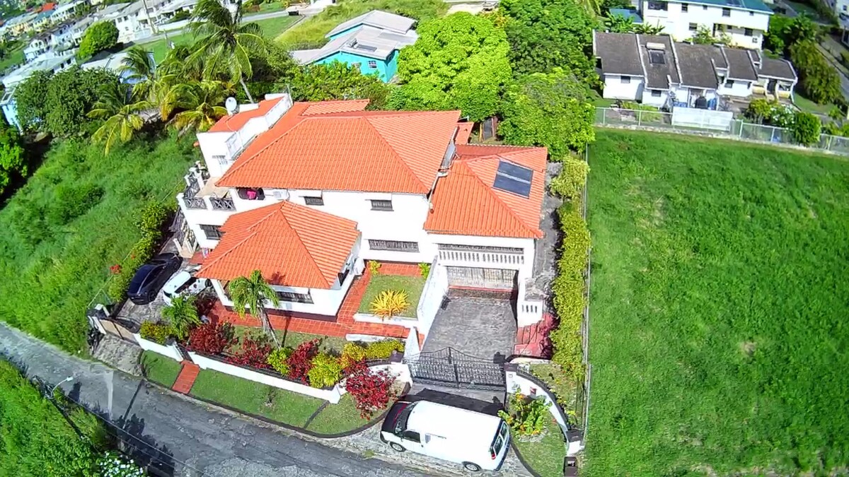 The spacious property features a two-story house with a red-tiled roof and lush greenery surrounding it. A well-maintained garden showcases colorful plants, while a large pool can be seen in the background, offering a backdrop of serenity amidst the landscape.