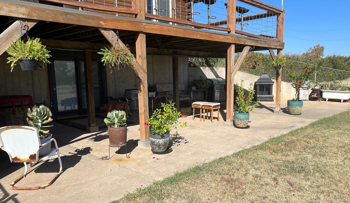 A shaded outdoor patio area is situated beneath a wooden deck, featuring a small table and various potted plants. Several chairs are arranged for seating, while a grill and additional outdoor furniture are present against the wall.