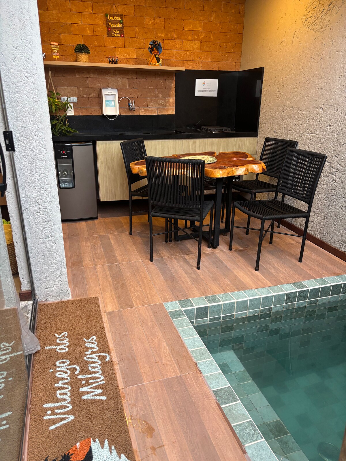 An outdoor dining area is shown, featuring a wooden table surrounded by six black chairs. A compact kitchenette is visible, equipped with a refrigerator and kitchen essentials. A private pool with a tiled border adds a refreshing element to the space, set against a simplistic wall backdrop.
