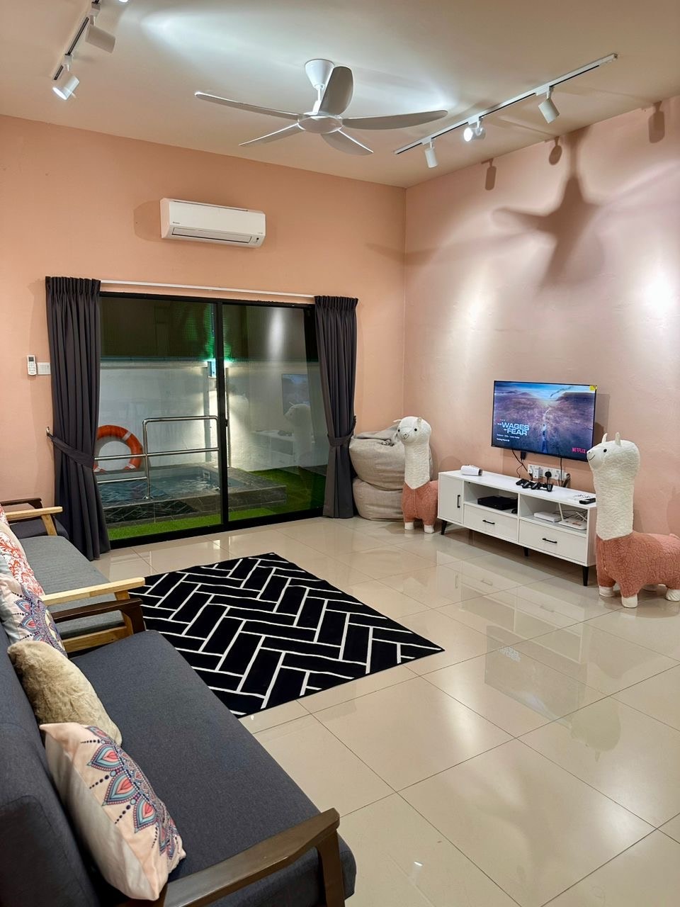 A cozy living area features a dark sofa adorned with patterned cushions, complemented by a decorative rug. A large sliding glass door opens to an outdoor space, while two charming llama figurines add a playful touch. A wall-mounted TV sits opposite the seating area.
