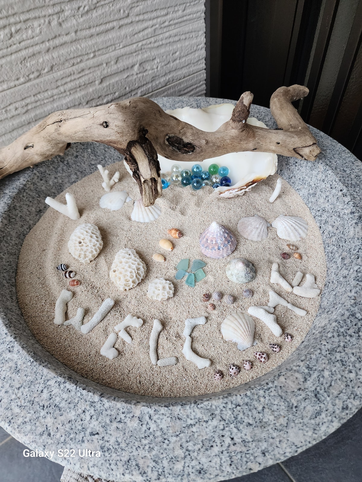 A decorative welcome display featuring a variety of shells and stones arranged in a sandy bowl. The word 'WELCOME' is emphasized with smaller shells, surrounded by larger shells and colored stones, creating an inviting atmosphere.