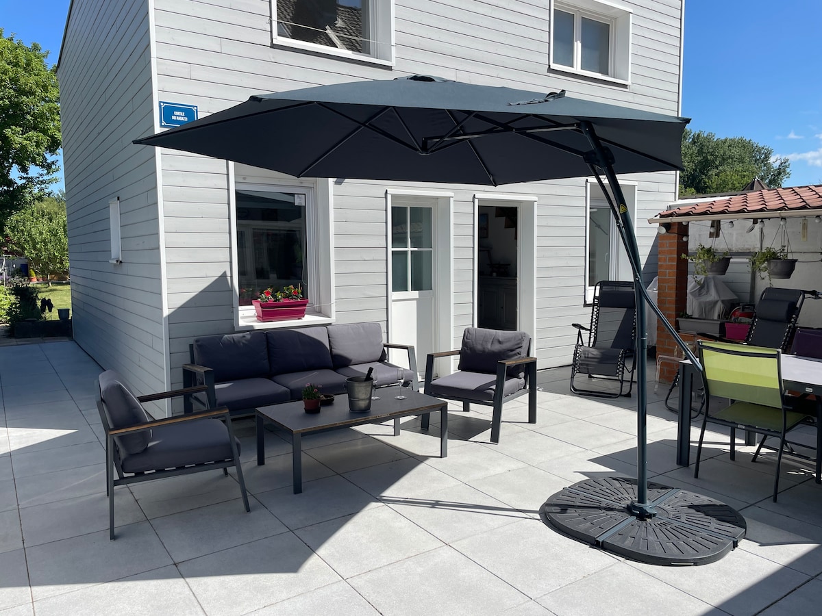An outdoor seating area is presented with a large umbrella providing shade. Comfortable seating includes a sofa and chairs arranged around a coffee table, with vibrant planters visible at window sills, and additional seating in the background.