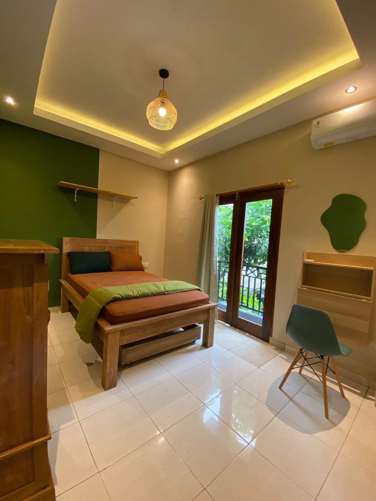 The room features a spacious bed with earthy-toned bedding against a two-tone wall. A small desk with a chair is present, along with a wardrobe. Natural light enters through glass doors that open to a mini balcony, surrounded by greenery.