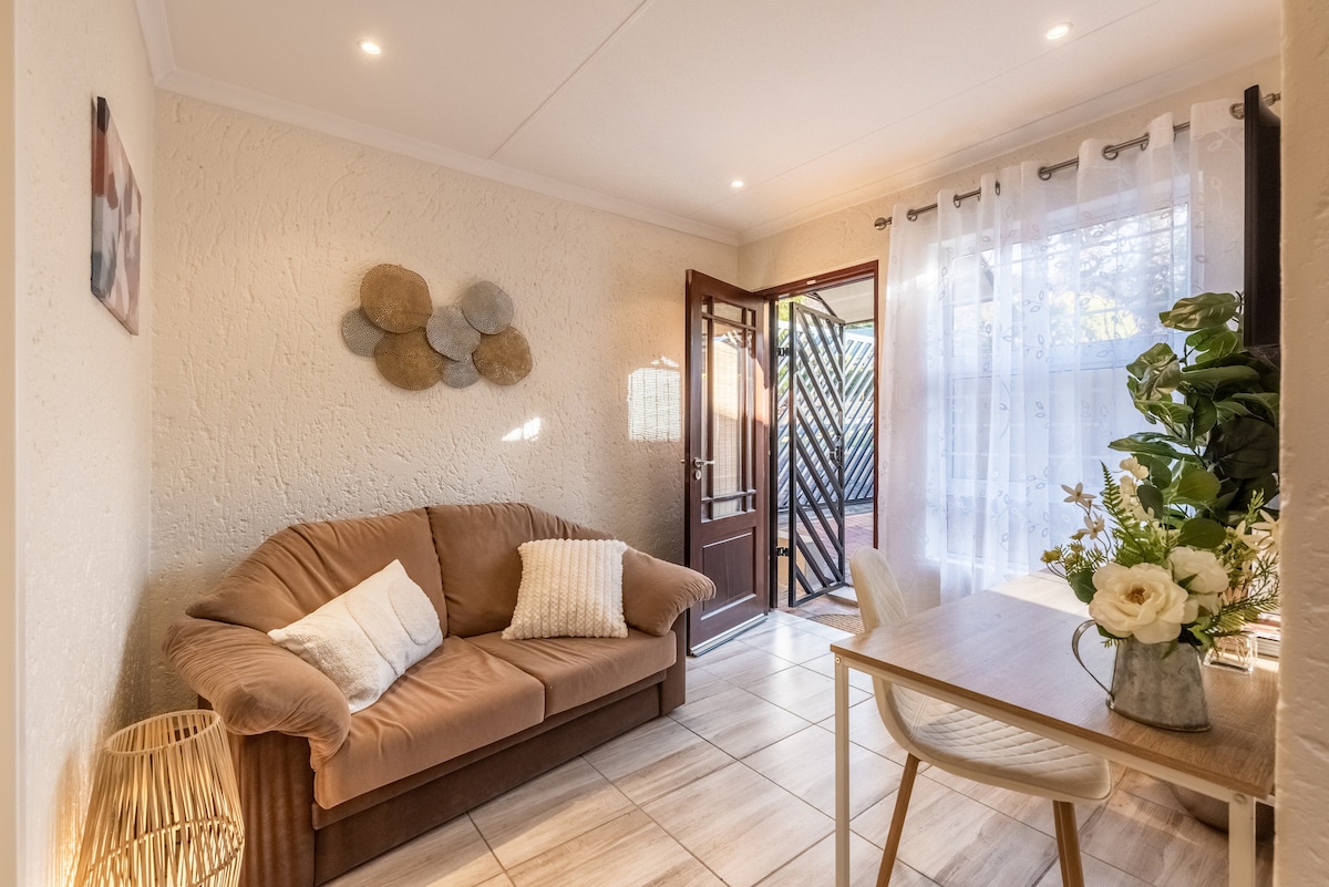 A cozy seating area is presented with a soft brown couch and a light wooden table. Natural light enters through sheer curtains, illuminating the textured walls. A decorative wall piece and a small plant enhance the space's inviting feel, with a door leading to an outdoor area.