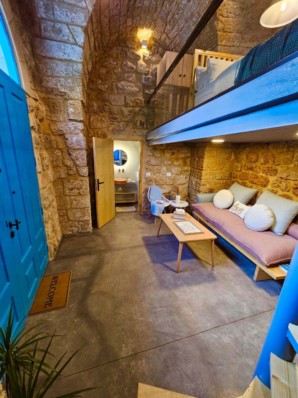 A stone-walled interior features a cozy seating area with a light sofa and a coffee table, complemented by decorative cushions. A modern bathroom is visible through an open door. The wooden door with vibrant blue paint adds contrast, while soft lighting enhances the welcoming atmosphere.