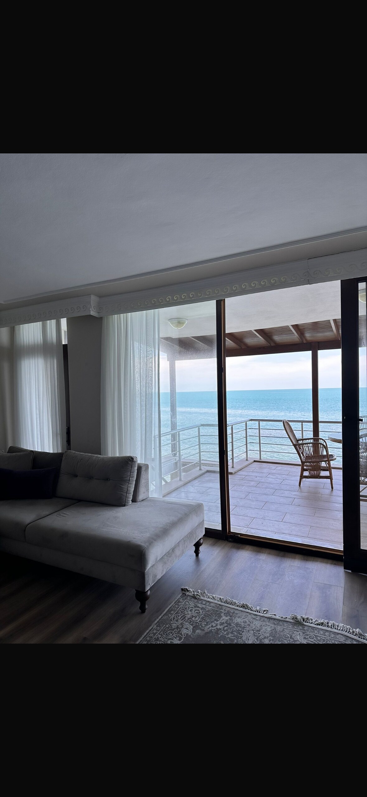 A bright living area features a comfortable gray sofa, with large glass doors that open to a spacious balcony overlooking the sea. Sheer white curtains gently diffuse the natural light, while a wooden chair invites relaxation on the outdoor area.
