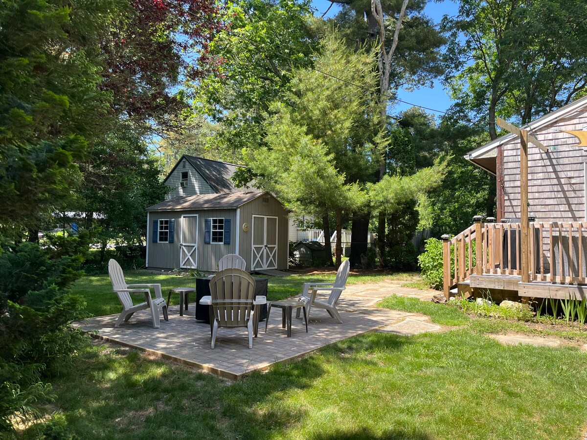 An outdoor seating area features a circular arrangement of Adirondack chairs surrounding a stone fire pit. Lush green trees provide shade, while a cozy shed is visible in the background. A path leads to a deck area beside a separate building.