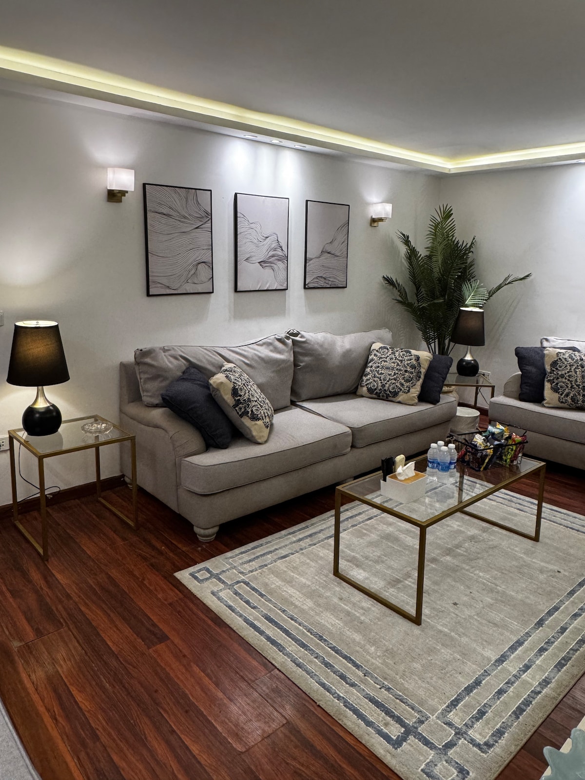 A living area is presented with two neutral-toned sofas adorned with decorative throw pillows. A low coffee table sits at the center, arranged with various items, including snacks and a welcome package. Subtle wall art adds character, while soft lighting highlights the space.