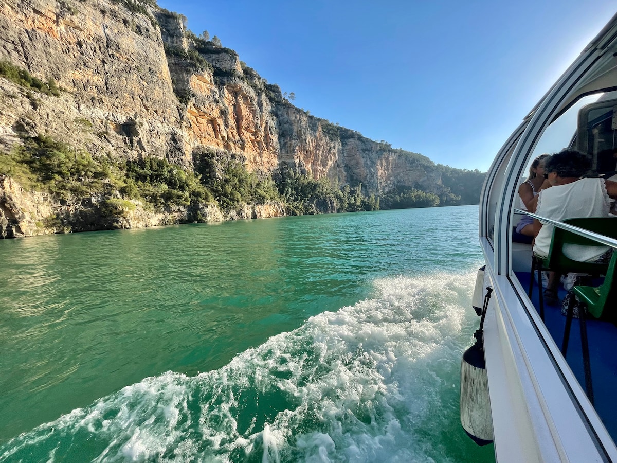 A boat is seen navigating through turquoise waters alongside rugged cliffs. Lush vegetation is present on the rocky terrain, creating a vivid contrast with the clear sky above. Passengers are seated inside, enjoying the scenic views outside the large windows.