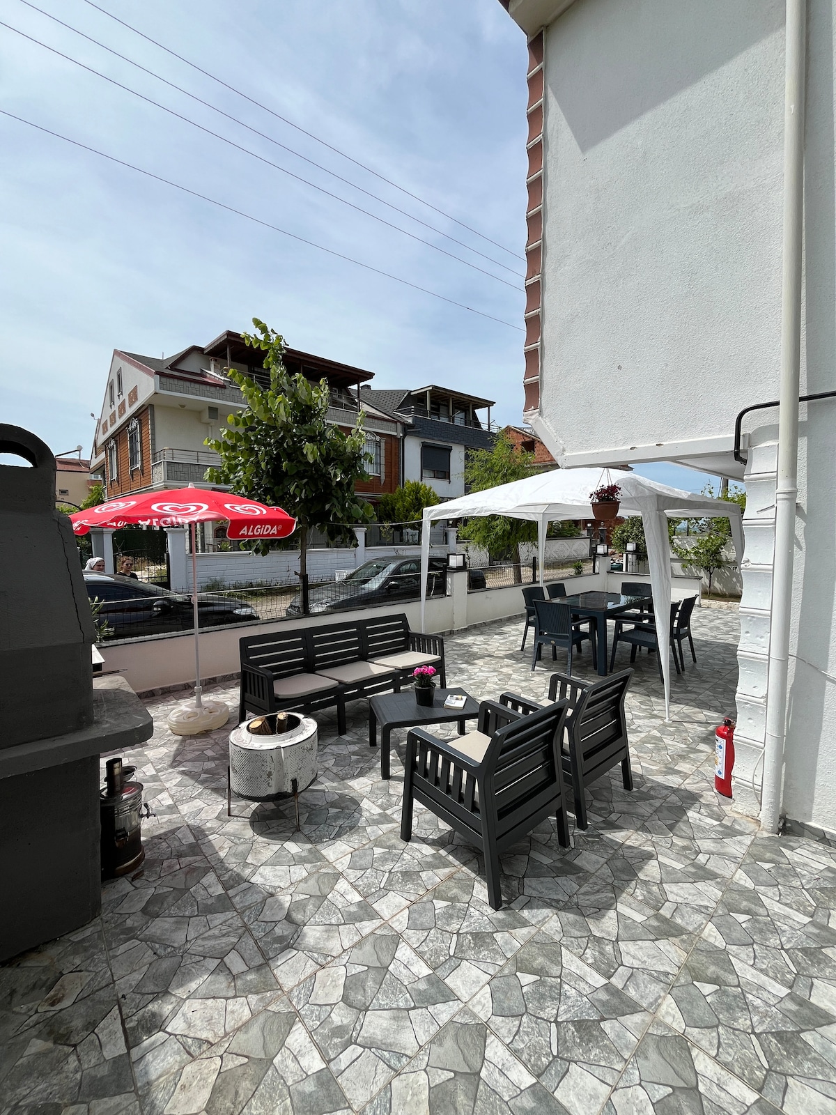 An outdoor terrace area features multiple seating arrangements with black metal chairs and tables. A shaded area with a white canopy offers a comfortable spot, while a grill is positioned nearby. The ground is paved with patterned tiles, adding texture to the space.