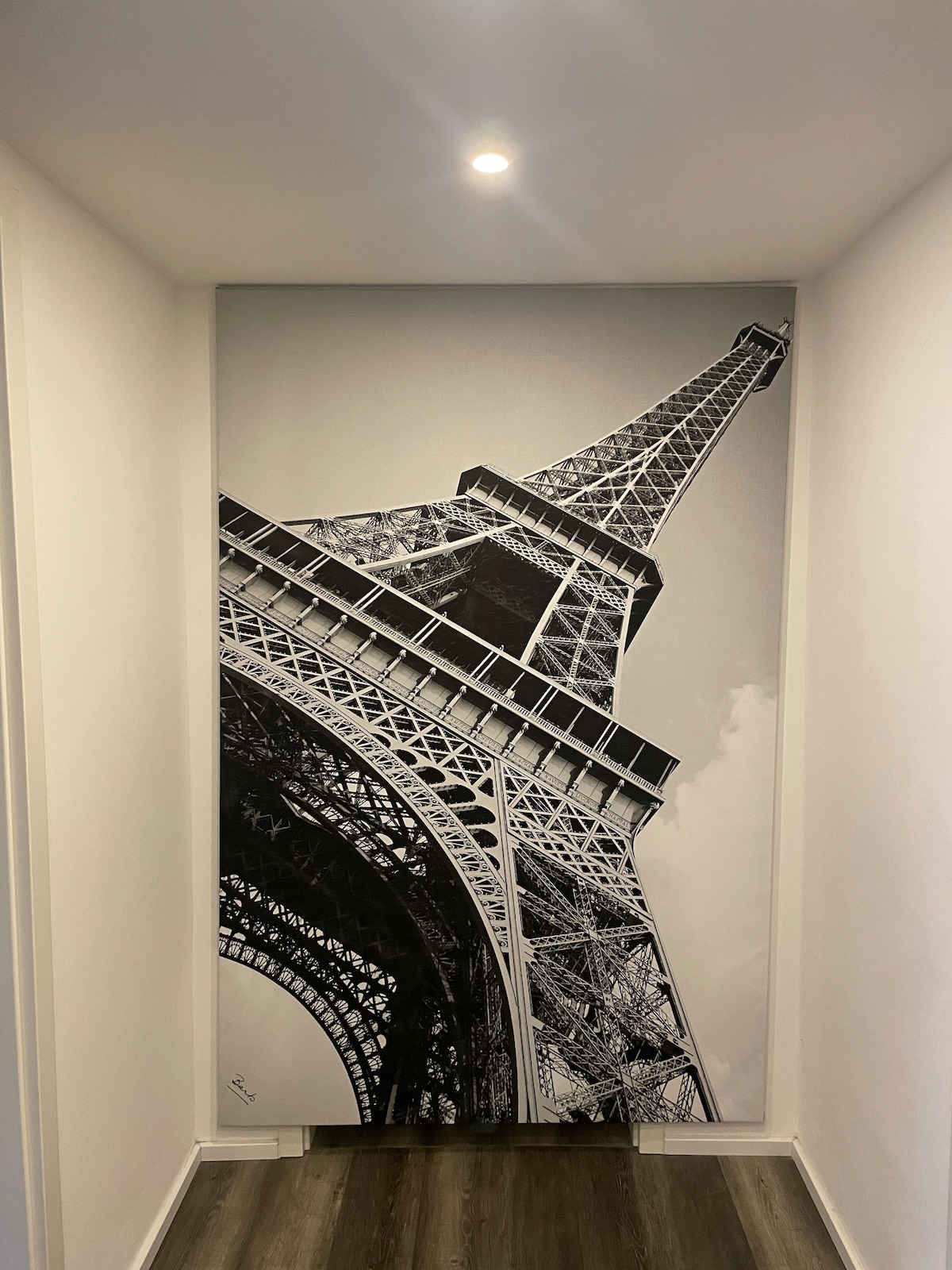 A striking monochrome mural of the Eiffel Tower covers one wall, positioned at an angle. The design emphasizes the tower's intricate ironwork and towering structure. Soft lighting from above highlights the smooth white walls and wooden flooring that complement the artwork.