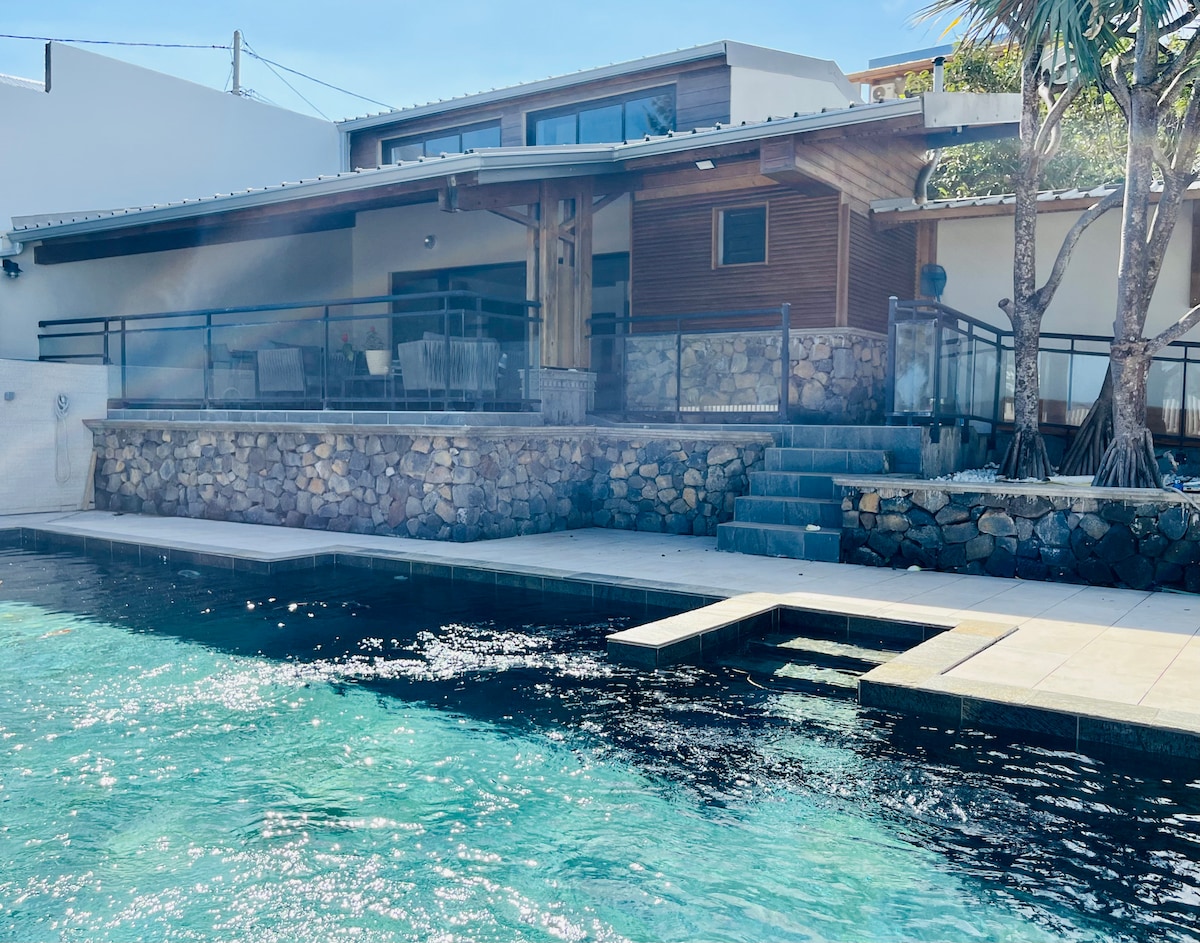 A modern home features a natural stone exterior with wooden accents. The structure overlooks a large heated swimming pool, complemented by a spacious terrace and steps leading to the water. Lush greenery is visible, enhancing the outdoor environment.