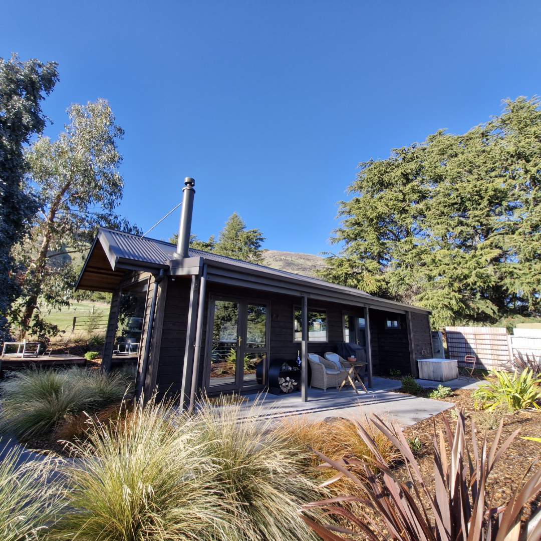 A country cabin is set amidst greenery, showcasing a modern exterior with large windows. A covered porch features seating options. Surrounding plants and trees enhance the serene atmosphere. The structure includes a chimney, indicating a wood burner for warmth.
